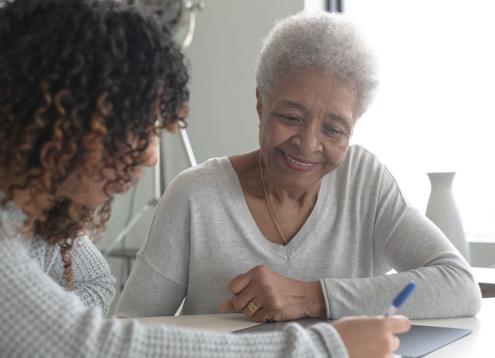 A woman in foreground is writing on paper. Elderly woman in background is smiling at her.